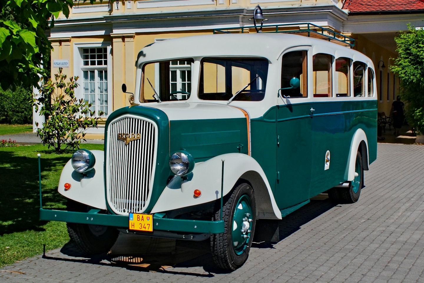 Vintážny tyrkysový a krémový autobus zaparkovaný pred žltou budovou.